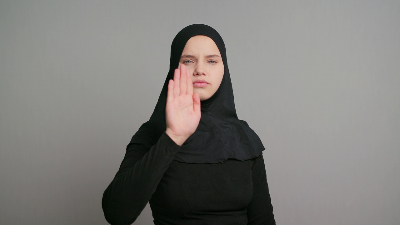 年轻的阿拉伯少女，戴着黑色头巾的穆斯林女学生愤怒地在灰色的背景上做着停止的手势。本空间视频素材
