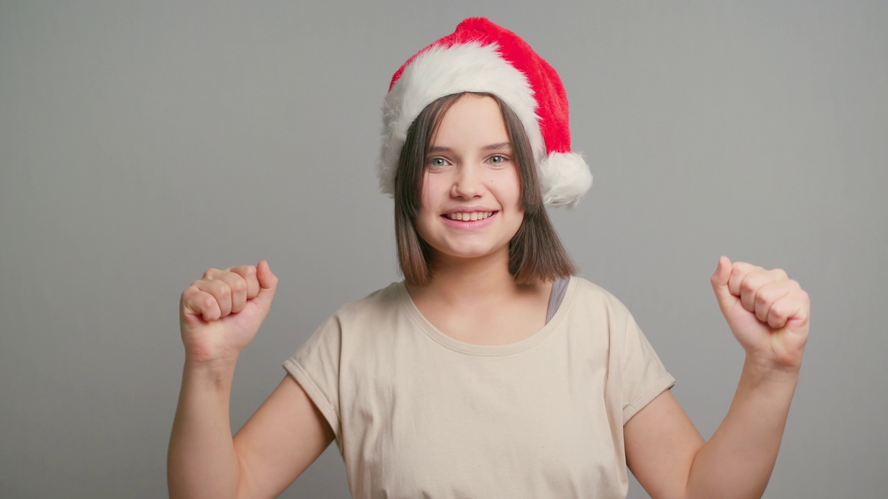 年轻的拉丁少女女学生戴着红色的圣诞老人帽子，快乐地孤立在灰色的背景上。本空间视频素材