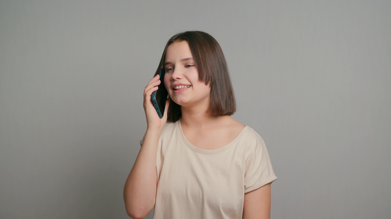 拉美裔少女微笑着快乐地拿着手机设备，用手机做电子商务网上购物，用手机上的应用程序站在灰色的背景上视频素材