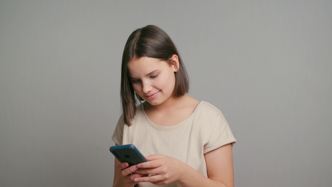 拉美裔少女微笑着快乐地拿着手机设备，用手机做电子商务网上购物，用手机上的应用程序站在灰色的背景上视频素材