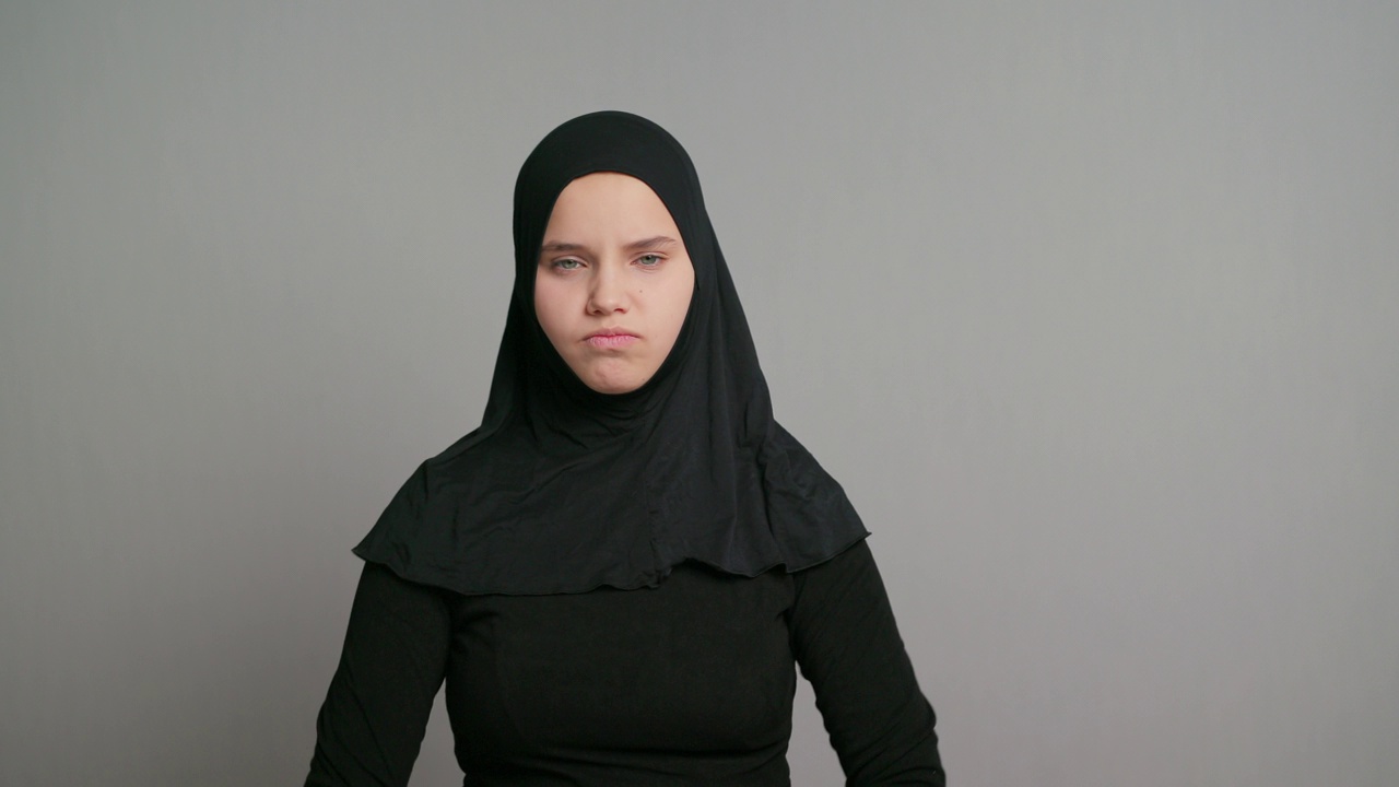 年轻的阿拉伯少女，戴着黑色头巾的穆斯林女学生，愤怒地交叉双手，孤立在灰色背景中。本空间视频素材