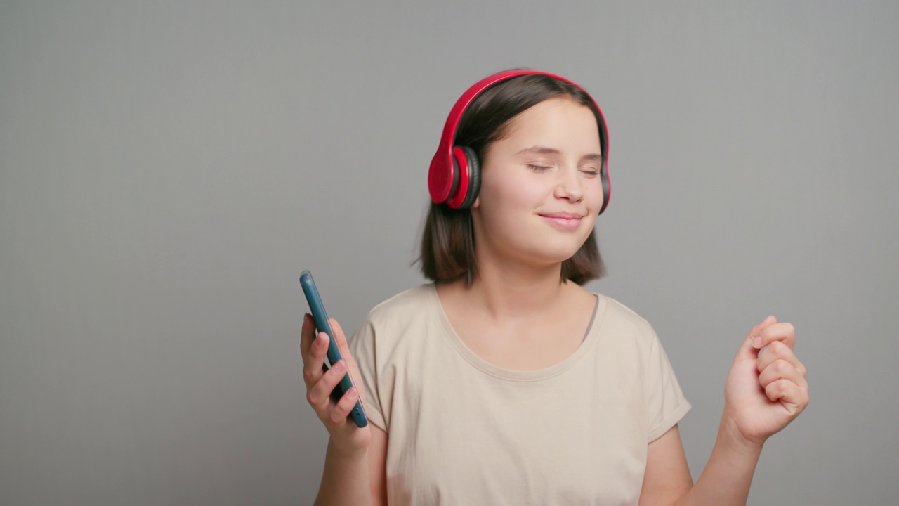 微笑的拉丁少女女子听播客电子书音乐歌曲歌手摇滚乐队戴着耳机耳机，选择音轨在手机孤立的灰色背景视频素材