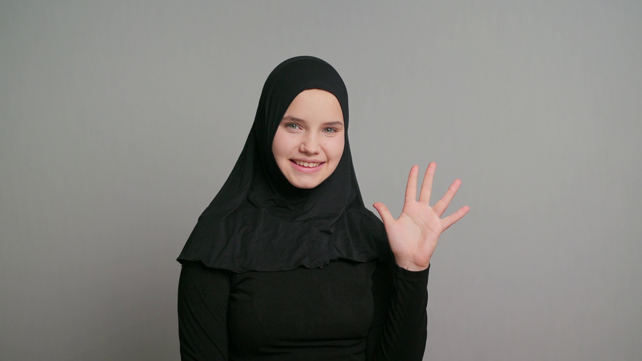 年轻的阿拉伯少女，戴着黑色头巾的穆斯林女学生，在灰色的背景上微笑着挥手打招呼。本空间视频素材