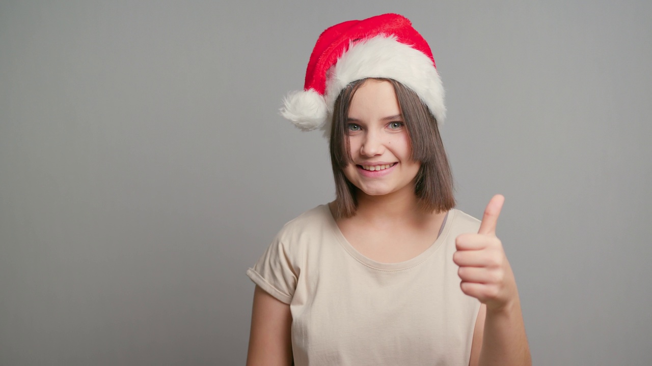 年轻的拉丁少女女学生戴着红色的圣诞老人帽，在灰色的背景上孤独地微笑着。本空间视频素材