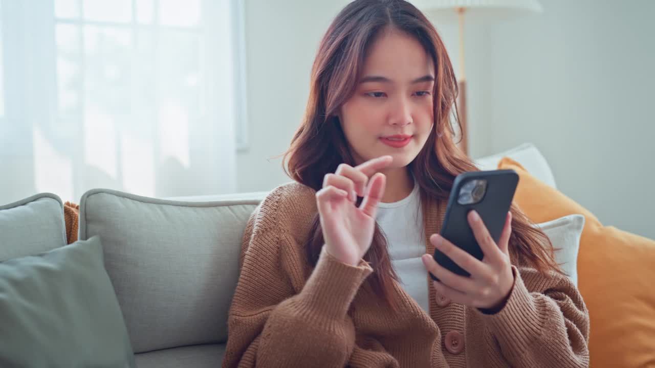 快乐的年轻亚洲女性在手机上冲浪社交媒体，享受惊人的新闻，女性坐在家里的沙发上玩游戏聊天社交媒体视频素材