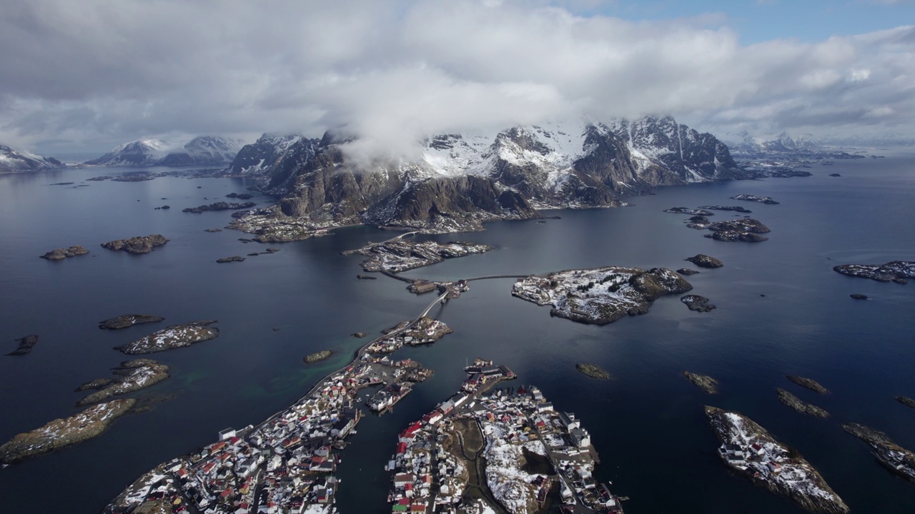 Henningsvær上空的无人机镜头，在云雾缭绕的天空下，展示了雪山、充满活力的蓝色水域和罗弗敦群岛分散的岛屿的惊人对比视频素材