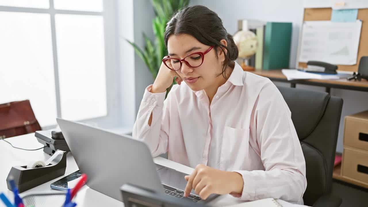 一位戴着眼镜的西班牙裔女士在井然有序的办公环境中聚精会神地在笔记本电脑前工作。视频素材