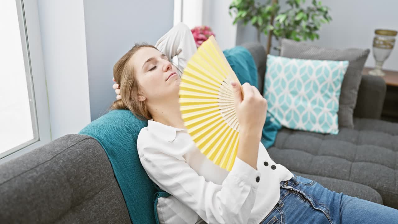 一名年轻女子躺在沙发上放松，在室内给自己扇风，营造出一种温暖、舒适的家庭氛围。视频素材