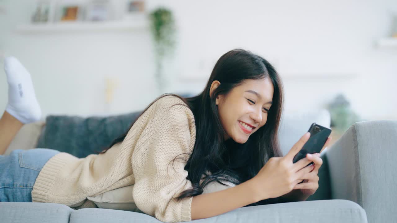 快乐的年轻亚洲女性在家里放松。女人笑着躺在沙发上，手里拿着手机。女孩用视频打电话给朋友视频素材