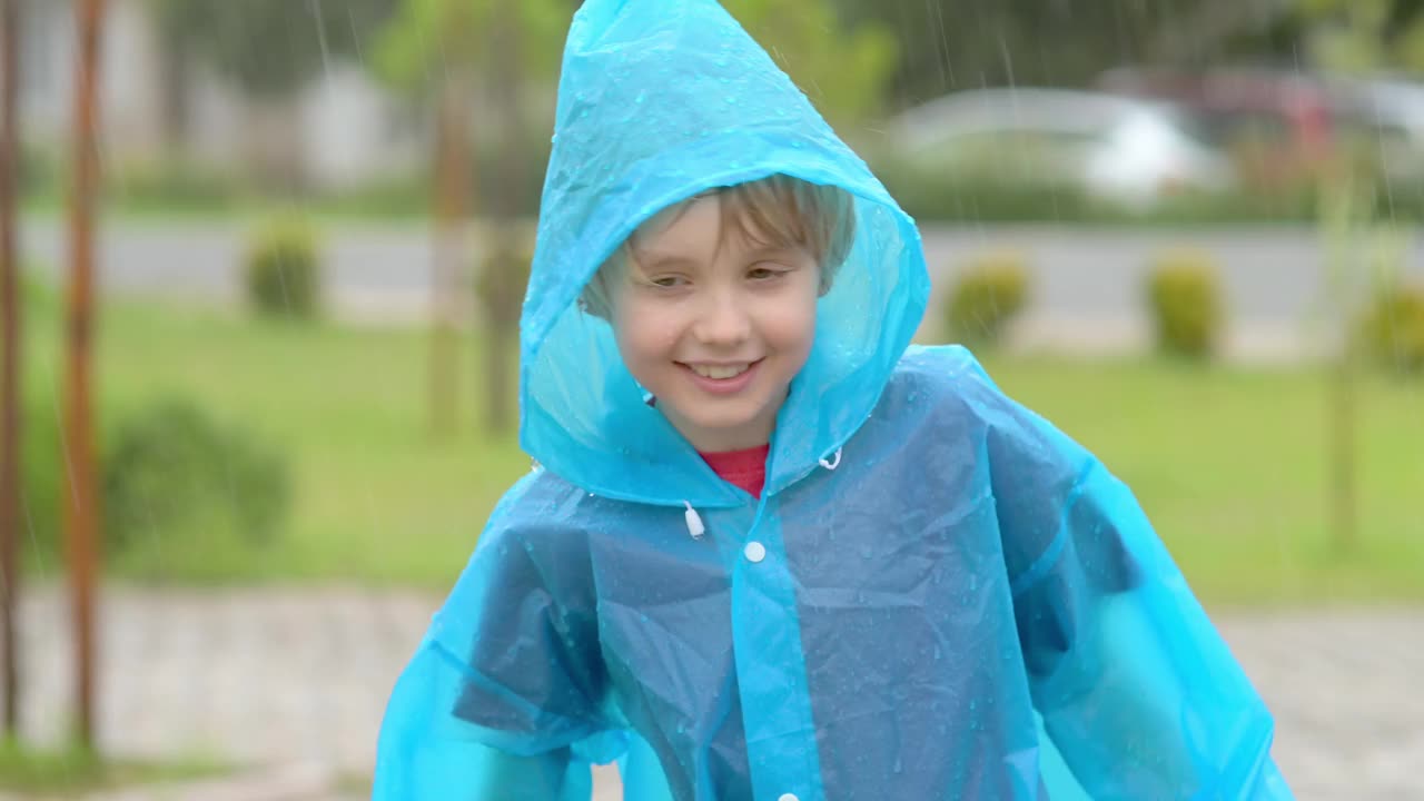 倾盆大雨中一个快乐的孩子的肖像。一个穿着蓝色雨衣的快乐男孩在倾盆大雨中走着，舌头上沾着水滴。视频下载