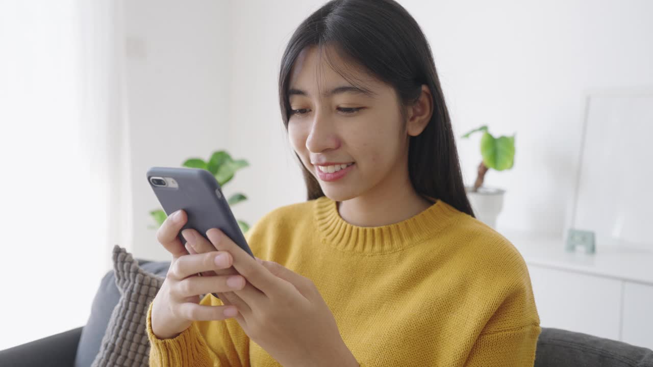 亚洲女孩在家用智能手机快乐地微笑打字视频素材