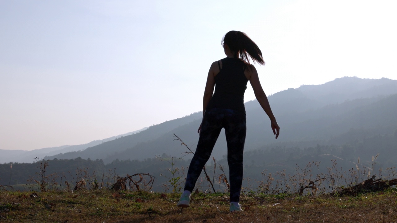 年轻女性在公园进行健身训练前的剪影。年轻女子在户外热身。她伸伸胳膊，望向别处。健康的生活方式和运动理念。视频素材