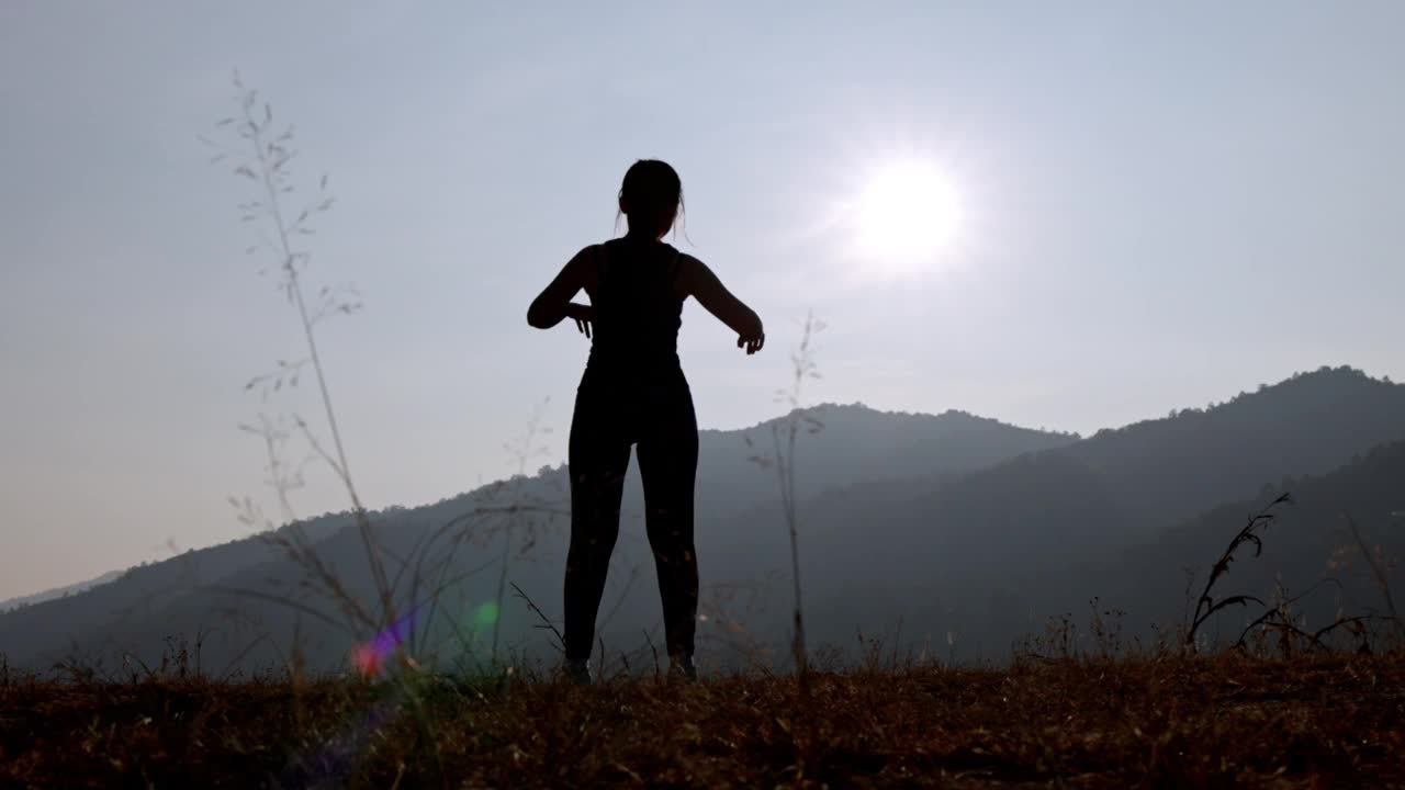 年轻女性在公园进行健身训练前的剪影。年轻女子在户外热身。她伸伸胳膊，望向别处。健康的生活方式和运动理念。视频素材