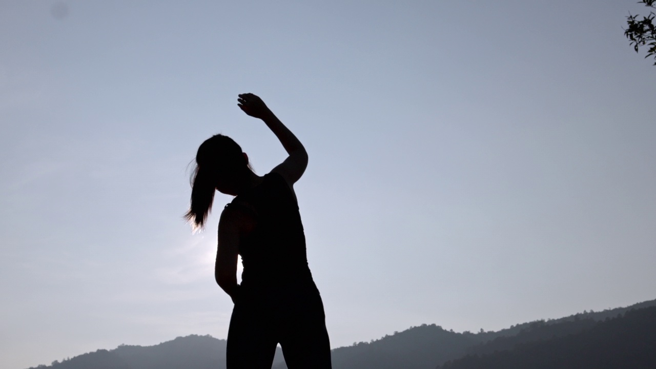 年轻女性在公园进行健身训练前的剪影。年轻女子在户外热身。她伸伸胳膊，望向别处。健康的生活方式和运动理念。视频下载