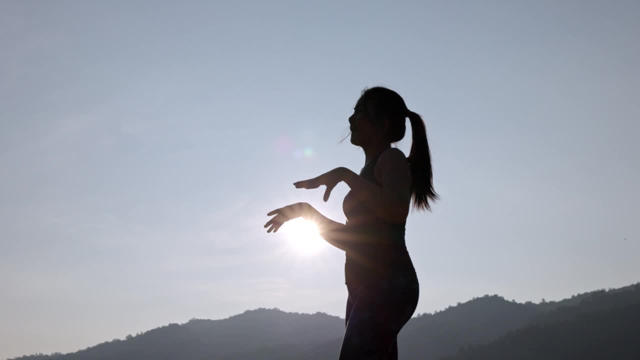 年轻女性在公园进行健身训练前的剪影。年轻女子在户外热身。她伸伸胳膊，望向别处。健康的生活方式和运动理念。视频下载