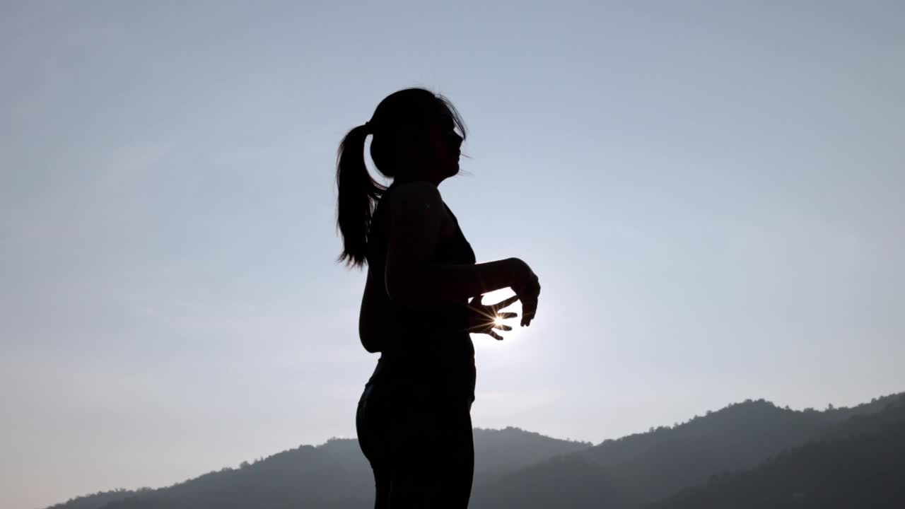 年轻女性在公园进行健身训练前的剪影。年轻女子在户外热身。她伸伸胳膊，望向别处。健康的生活方式和运动理念。视频下载