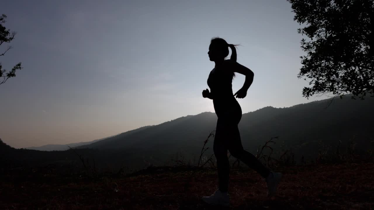 夕阳下身穿运动服的女子在山上慢跑的剪影。穿着运动胸罩在户外跑步的年轻亚洲女性。晚上锻炼身体。健康积极的生活方式理念。视频下载