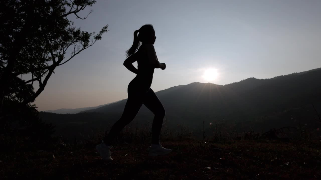 夕阳下身穿运动服的女子在山上慢跑的剪影。穿着运动胸罩在户外跑步的年轻亚洲女性。晚上锻炼身体。健康积极的生活方式理念。视频下载