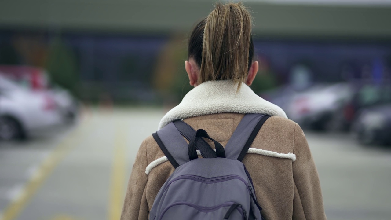 背着双肩包、梳着马尾辫的女旅行者推着大行李箱走在机场、火车站停车场，慢动作的电影镜头视频素材