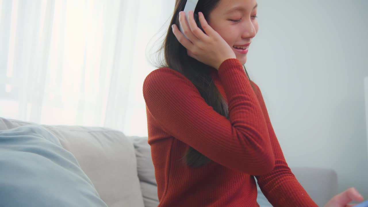 亚洲年轻女性在家里的客厅里拿着手机听音乐。迷人的漂亮女孩戴上数码耳机，在家里玩智能手机，感到快乐和放松视频素材