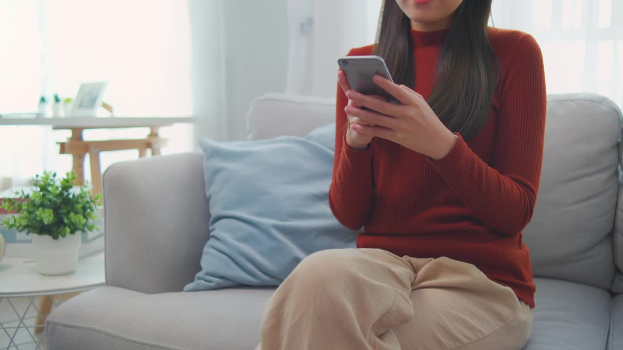 亚洲年轻女性在家里的客厅里拿着手机听音乐。迷人的漂亮女孩戴上数码耳机，在家里玩智能手机，感到快乐和放松视频素材