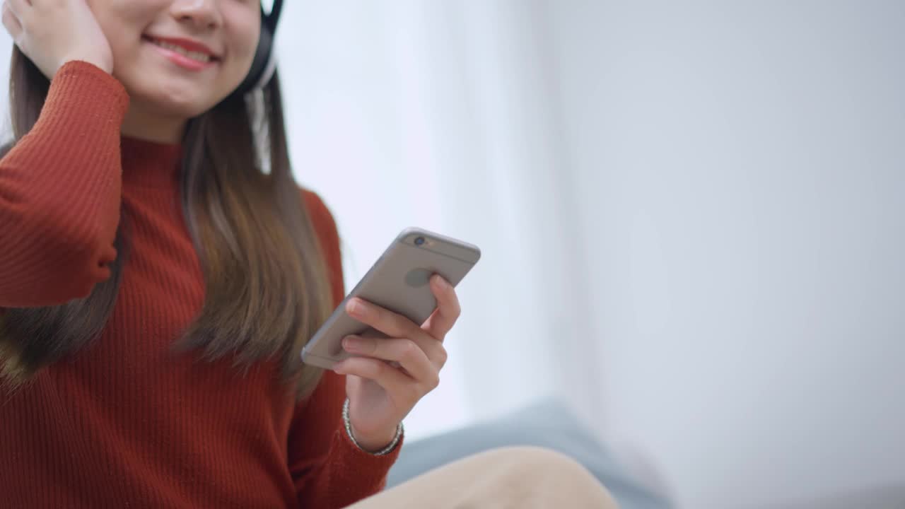 亚洲年轻女性在家里的客厅里拿着手机听音乐。迷人的漂亮女孩戴上数码耳机，在家里玩智能手机，感到快乐和放松视频素材