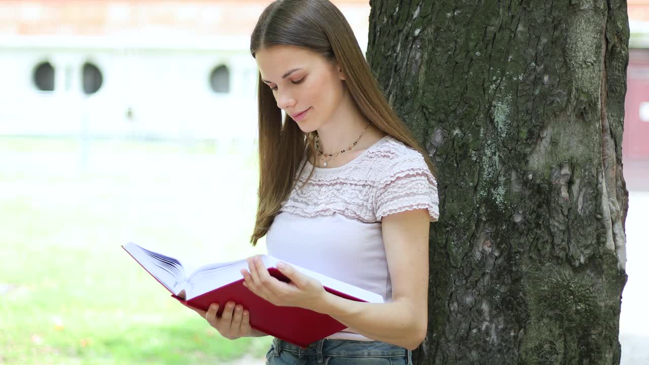 一个女学生在公园里靠在树上看书，面带微笑视频下载