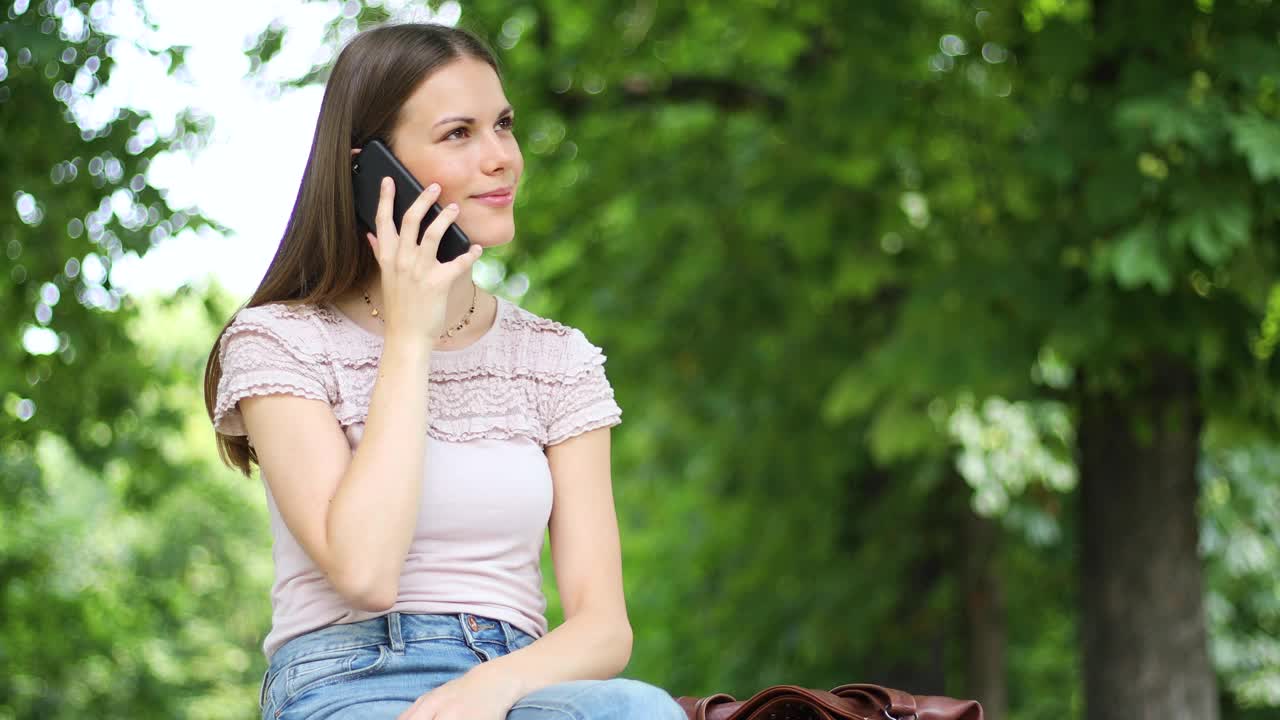 一个快乐的女人坐在长凳上，在户外打电话视频下载