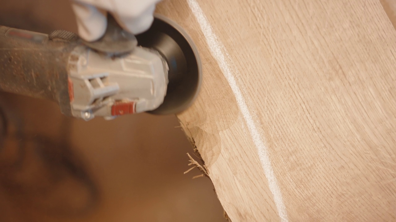 木工车间的木工过程的高角度视图，男工匠戴着防护手套用磨床打磨木板的角度，木材打磨后的车间地面上堆着木屑，工作台上的原木材料打磨视频素材