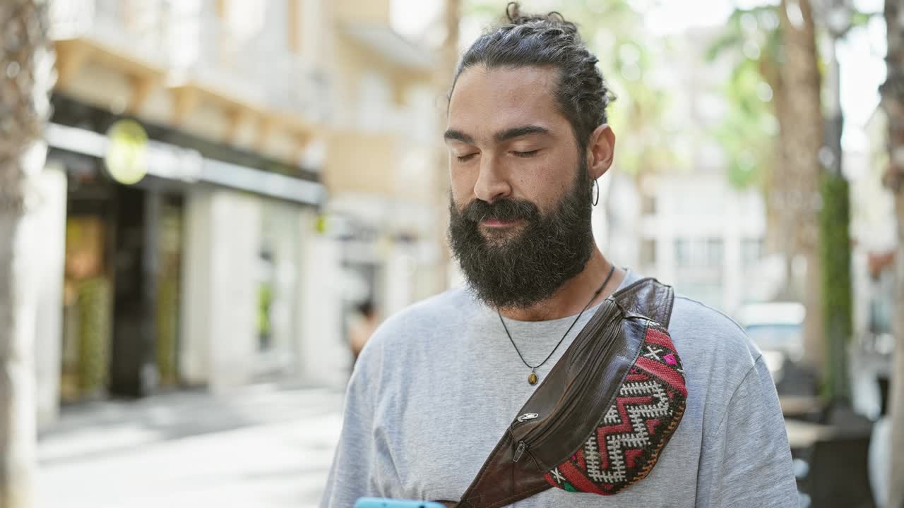 一位满脸笑容的大胡子男子一边走在城市街道上一边用智能手机说话，散发着城市生活方式和互联互通的气息。视频素材