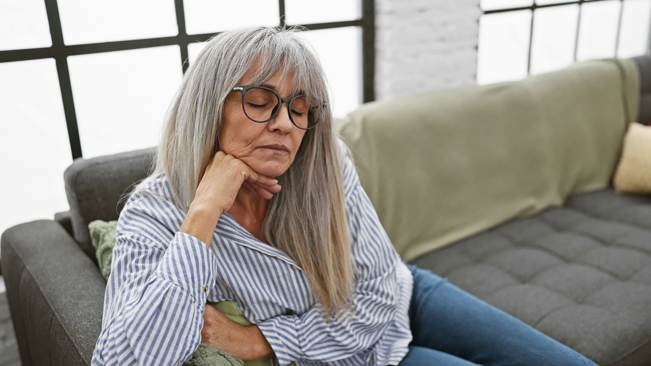 一个白发苍苍、戴着眼镜的成熟女人若有所思地坐在室内的沙发上视频素材