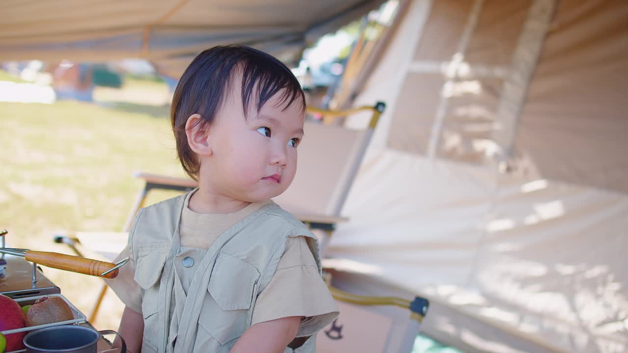 快乐的幼童和家人在露营地享受了一天，他穿着卡其色的衣服，背后是一个阳光普照的帐篷视频素材