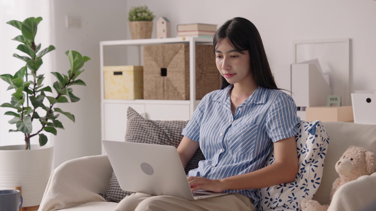 亚洲女孩坐在沙发上使用笔记本电脑，亚洲女人开心地微笑着在家里打字视频素材