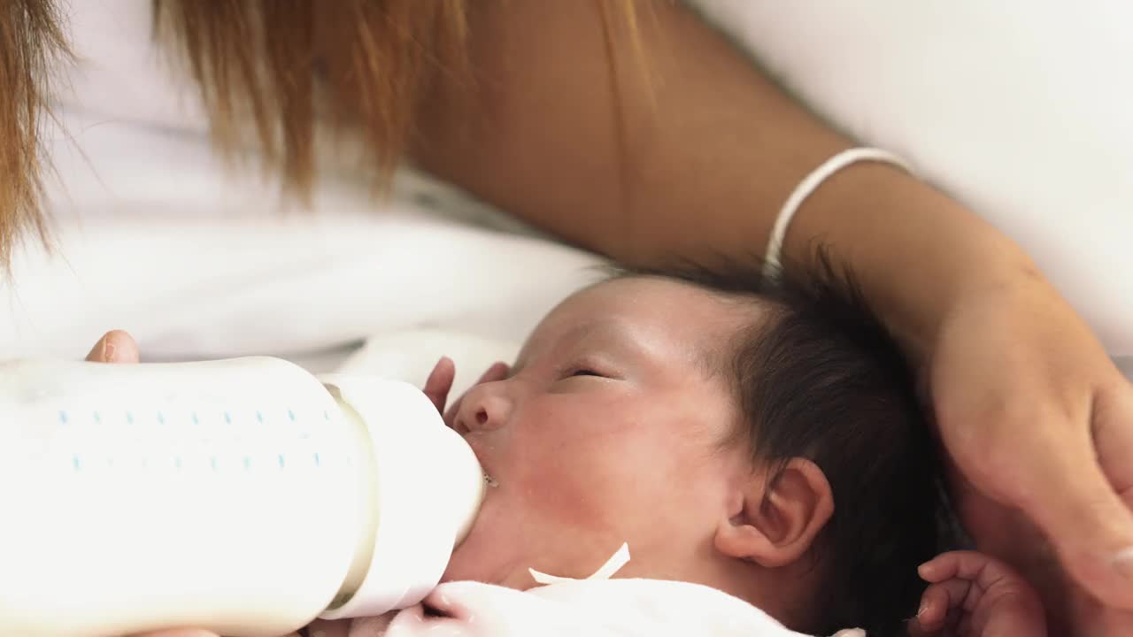 妈妈用爱给宝宝喂奶新生的宝宝脸从奶瓶里喝奶新生的女孩躺在床上感到困倦可爱的婴儿在家里用爱喂奶新生婴儿概念视频素材