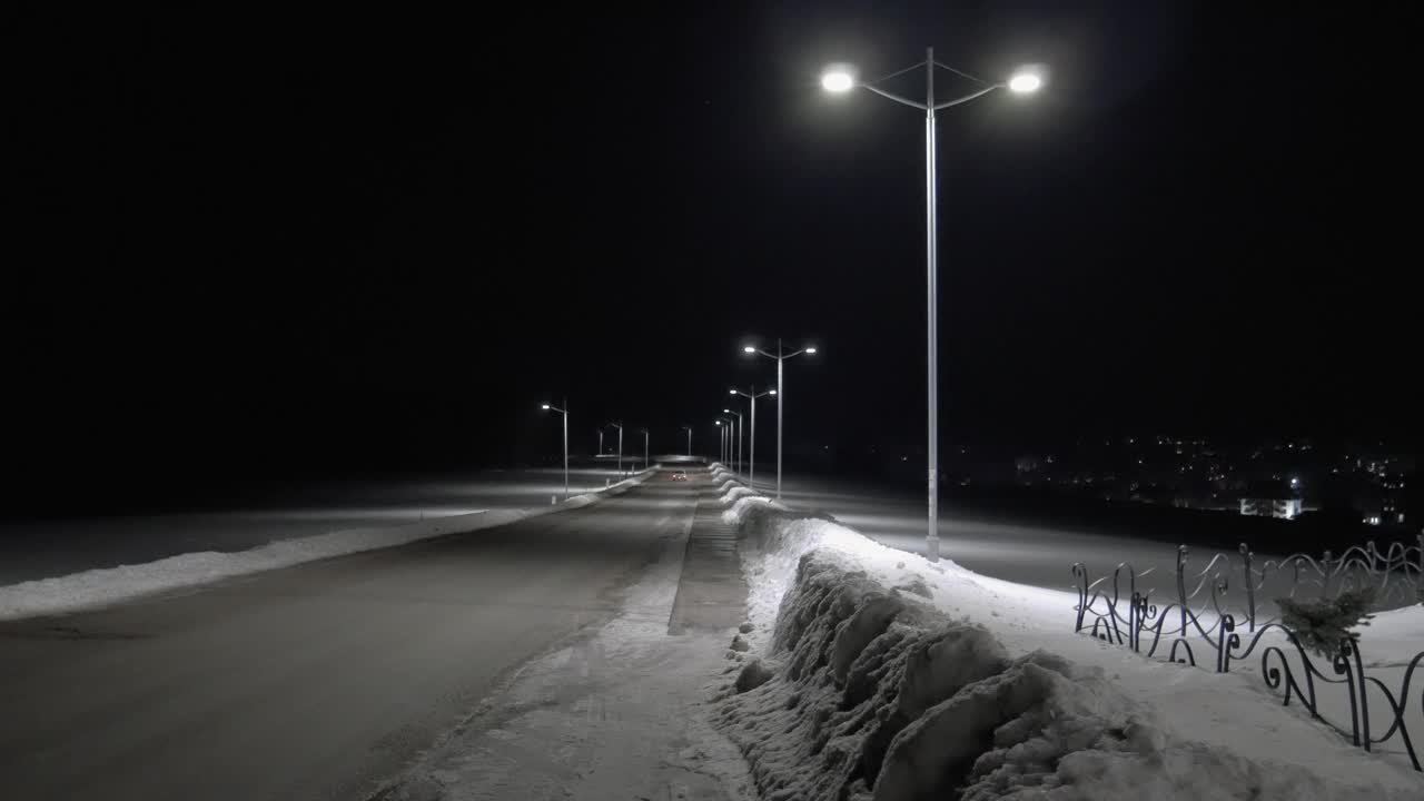 一辆小车在冬夜行驶在被雪覆盖的道路上。电线杆照亮道路。远处可以看到城市的灯光。手持拍摄的视频下载