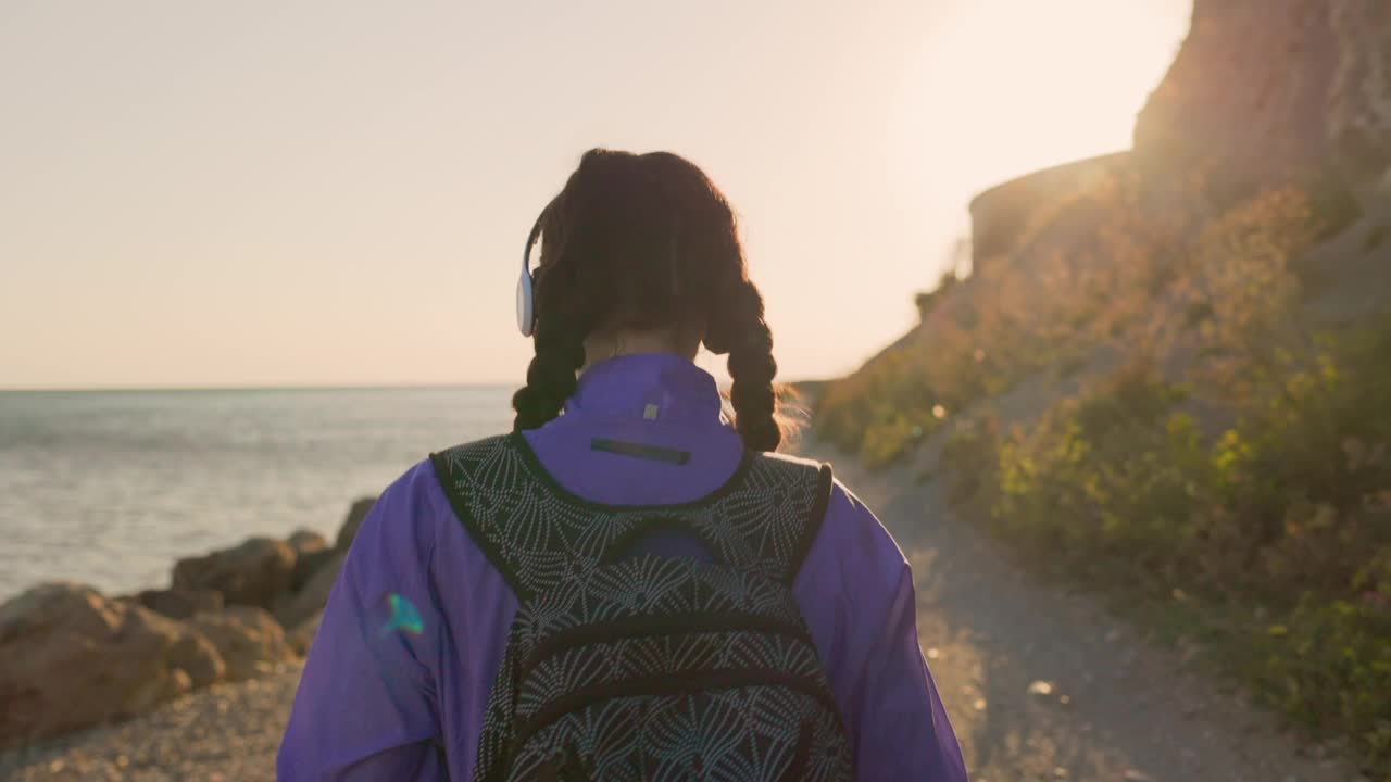 徒步旅行。一个扎着小辫子背着背包的年轻女子在海边散步，戴着耳机听音乐。孤独的心理学。自由和主动娱乐的概念视频下载