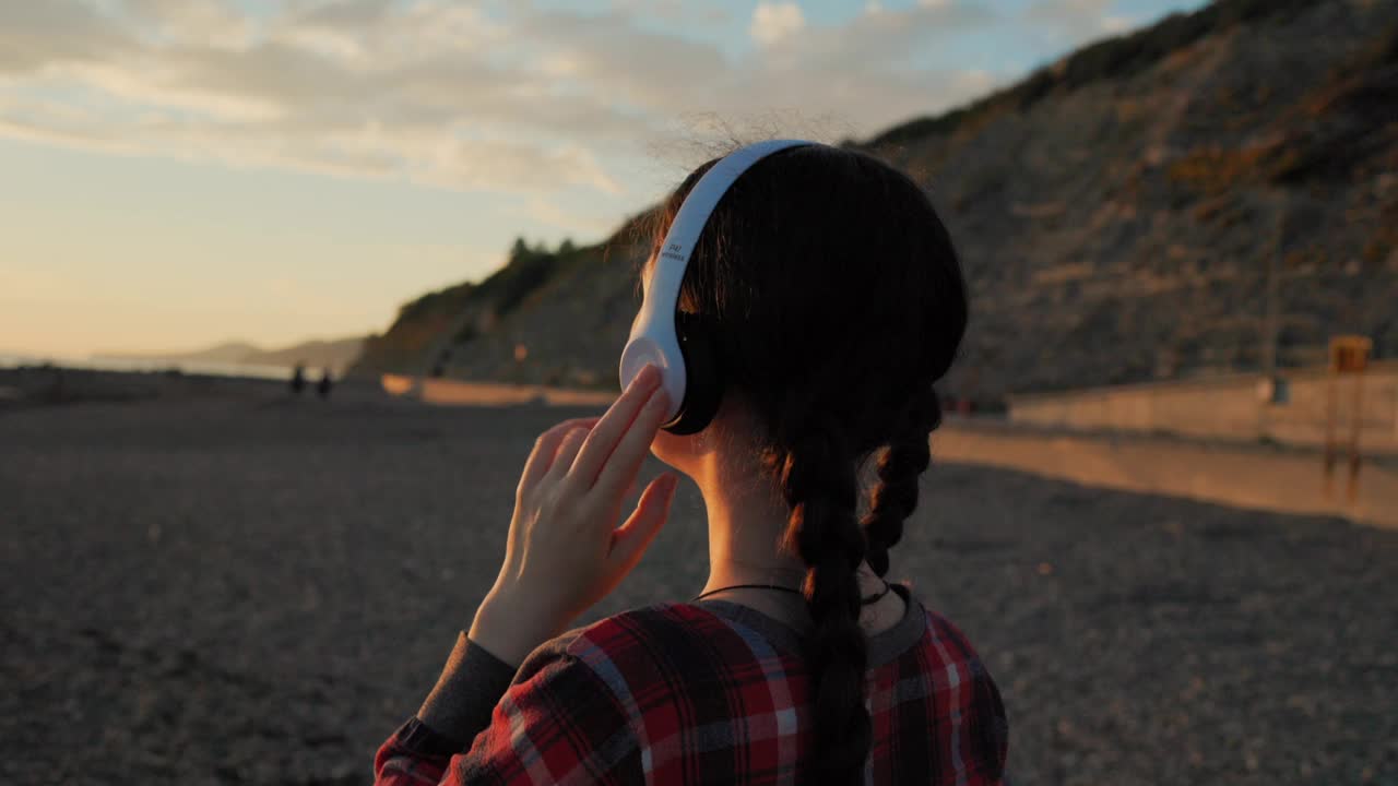 一个扎着小辫子的年轻白人女人，戴着耳机听音乐，看日落。背景是海滩和海洋。锅。自由和心理的概念视频素材