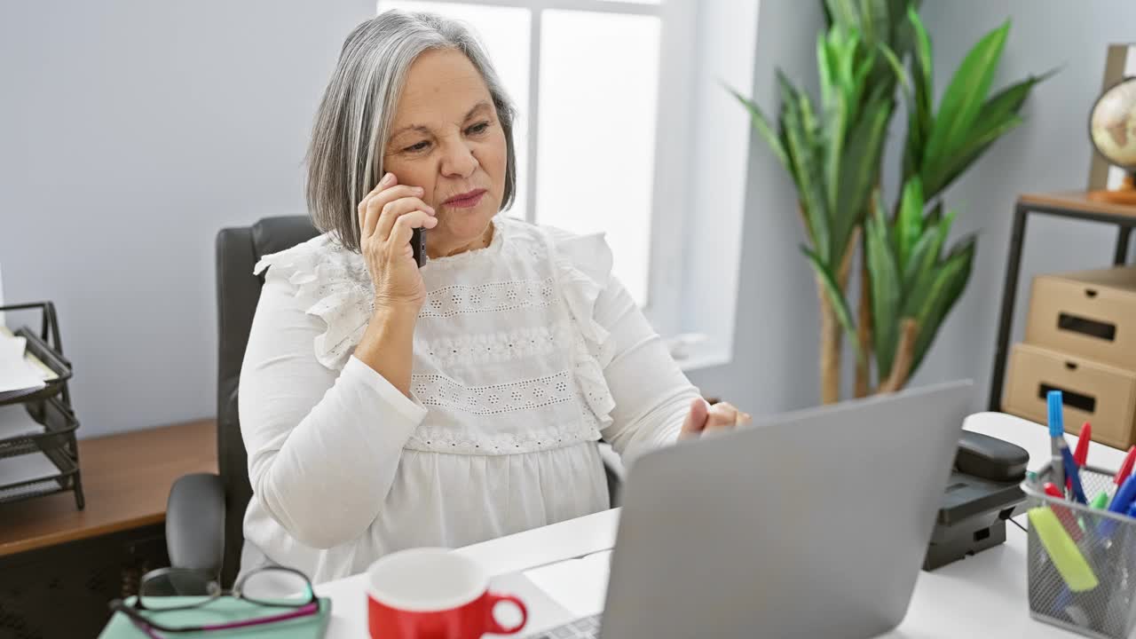 Senior woman conversing on phone at desk with laptop in office视频素材