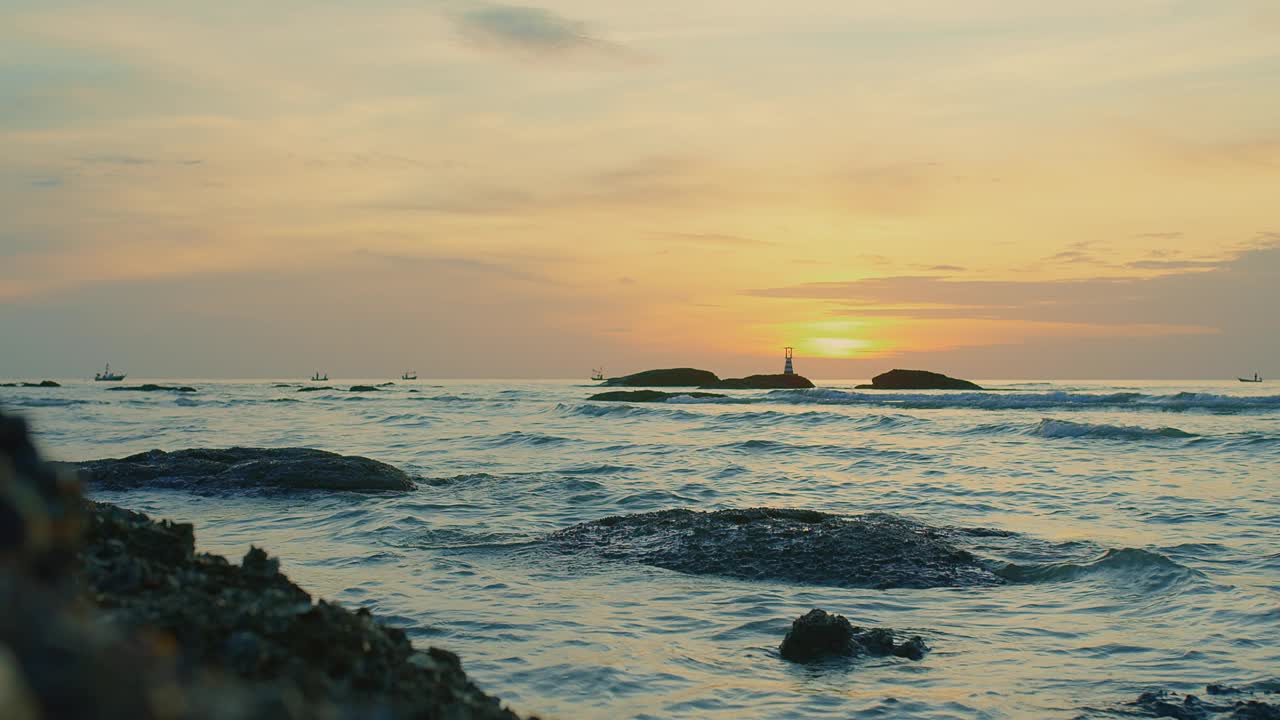 日落时宁静的海景，海浪轻轻地拍打着岩石，远处有灯塔的剪影视频素材