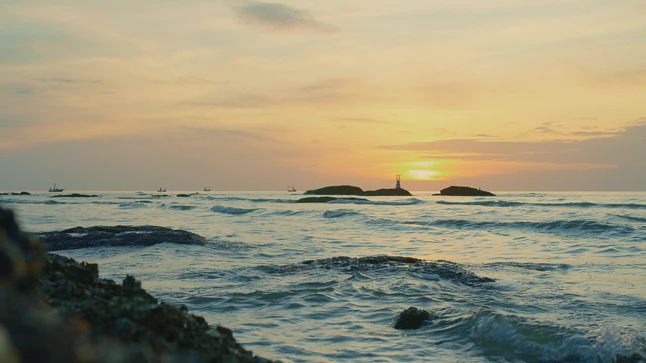 日落时宁静的海景，海浪轻轻地拍打着岩石，远处有灯塔的剪影视频素材