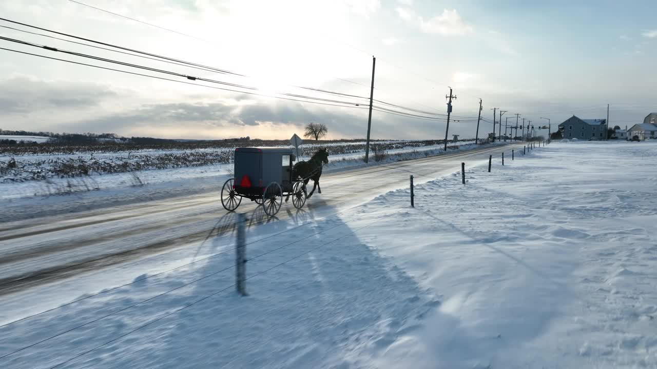 在夕阳的天空下，宾夕法尼亚州兰开斯特县下雪的道路上的阿米什马车。空中跟踪拍摄的雪在美国农村道路上漂移。视频素材