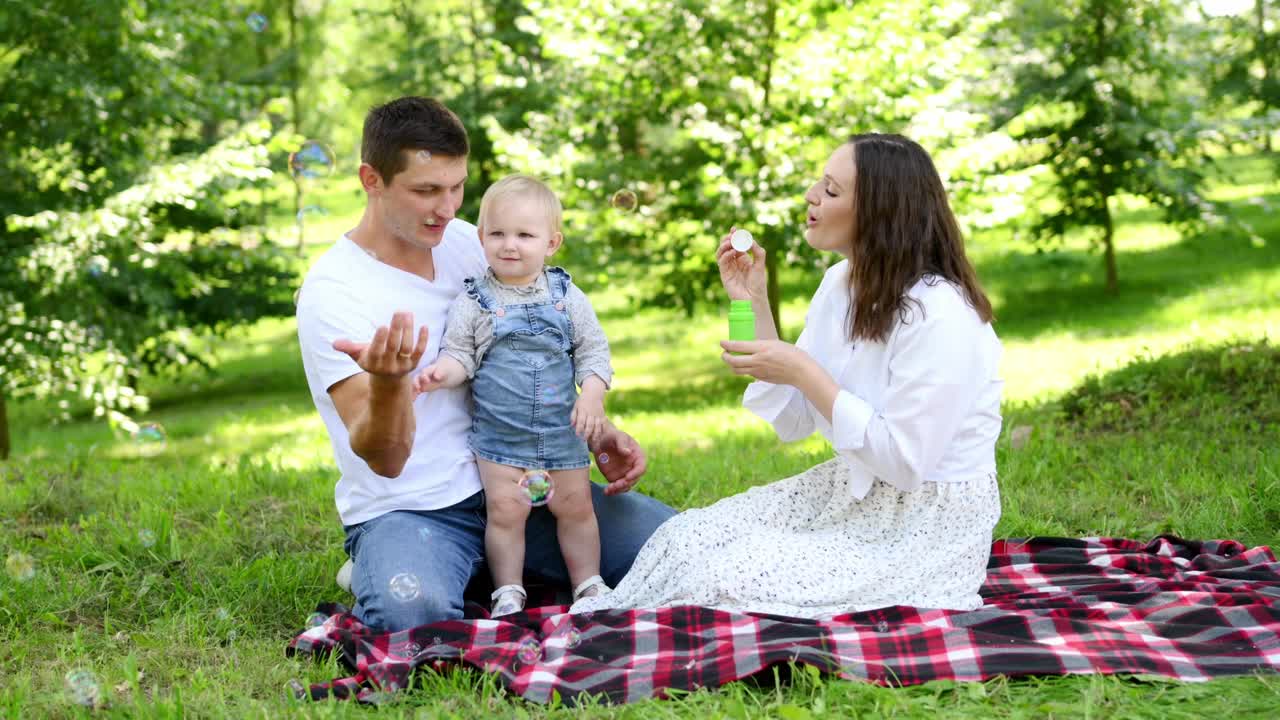 一个快乐的年轻家庭带着一个婴儿在公园里野餐，吹着肥皂泡。夏令时间概念。家庭日视频素材