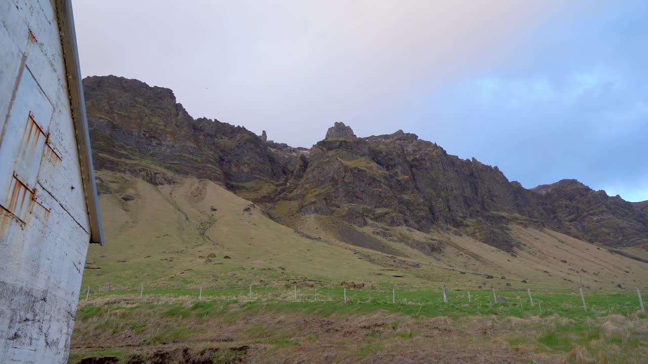 多云的天空下，崎岖的冰岛风景，绿色的草地和雄伟的山峰，乡村建筑的侧视图视频素材