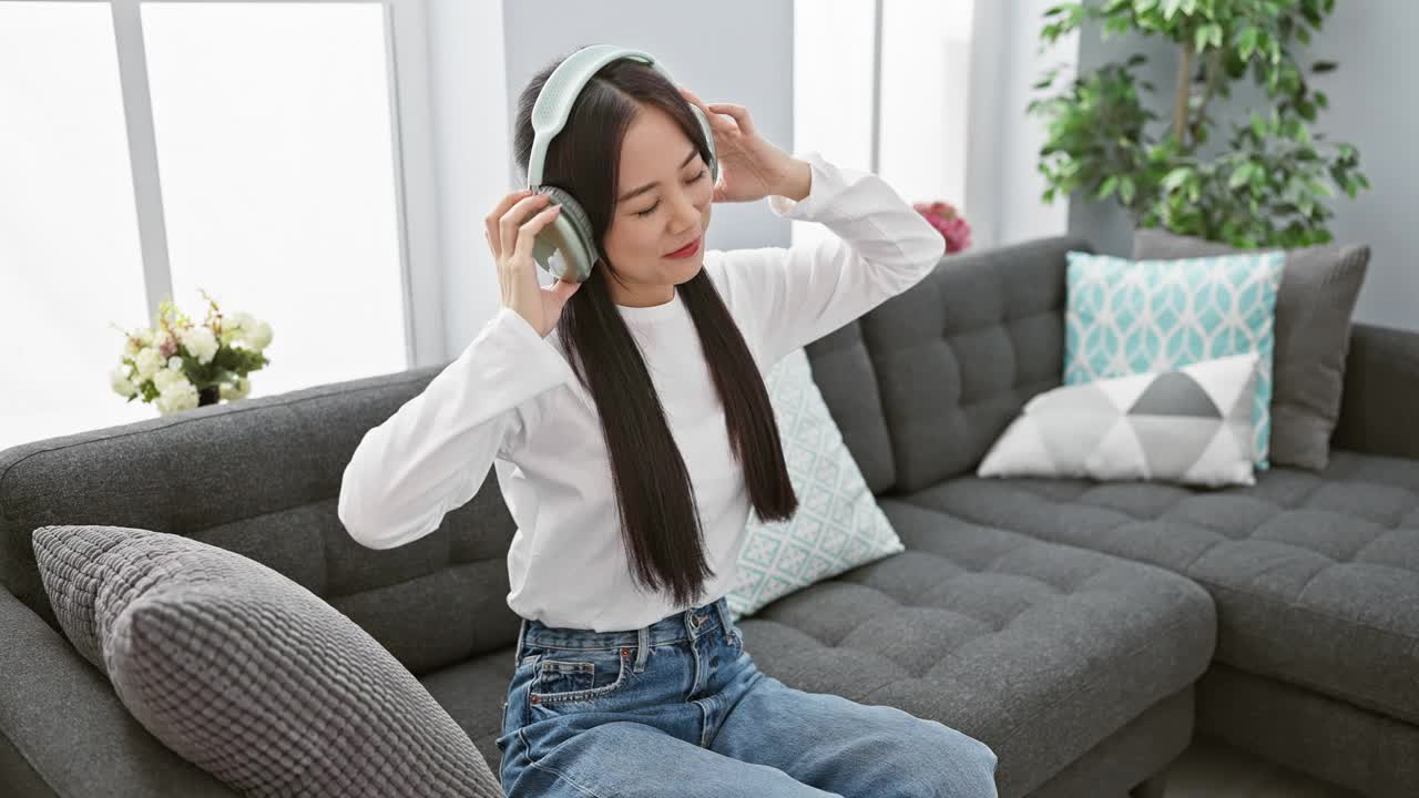 在明亮的客厅里，一位开朗的年轻亚洲女子坐在灰色的沙发上，戴着耳机欣赏音乐。视频素材