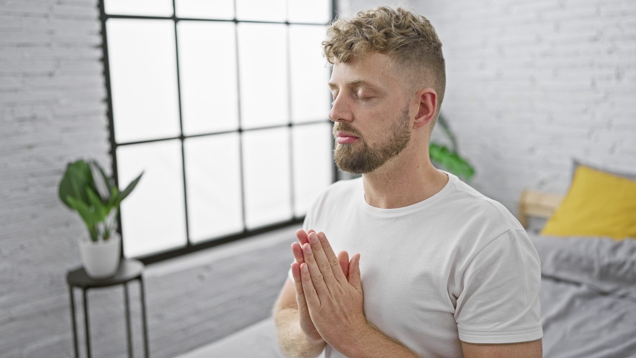 一个留着胡子的年轻白人男子在一间现代卧室里冥想，卧室有一扇大窗户，背景是一棵植物。视频素材