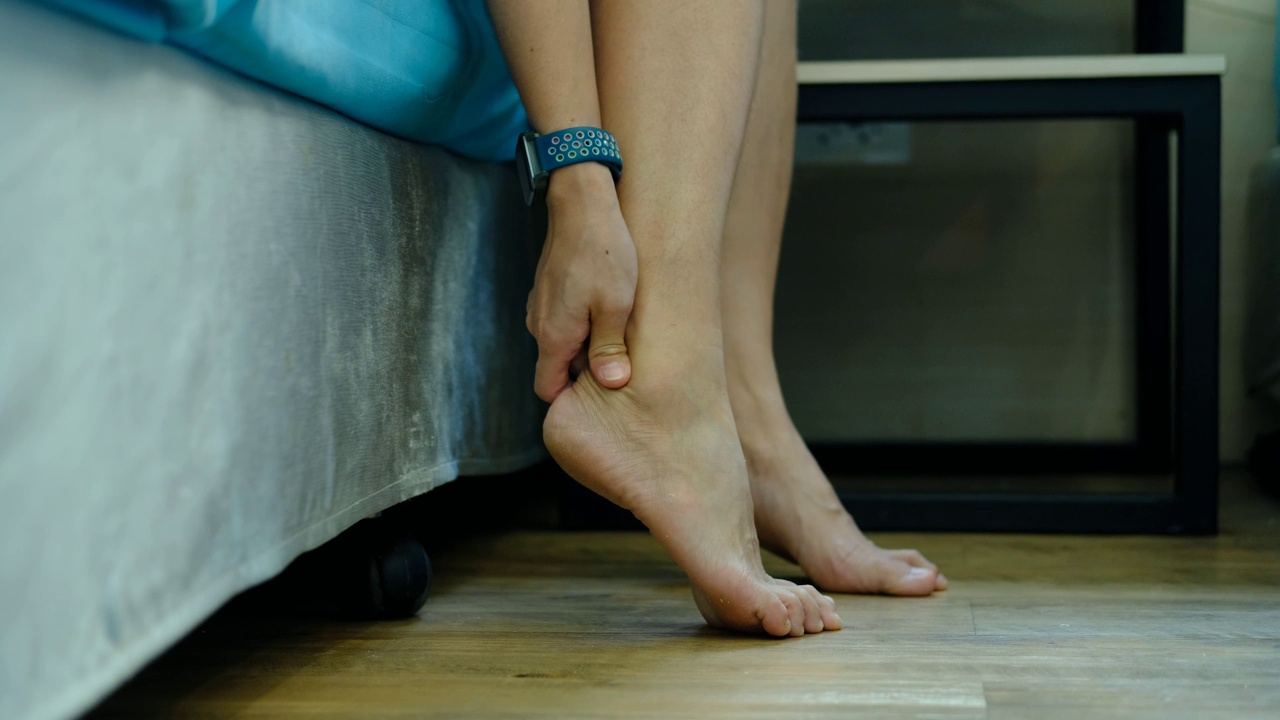 足部疼痛，亚洲女性坐在家里感觉足部疼痛，患有足部疼痛的女性用手按摩来放松脚底肌肉视频素材