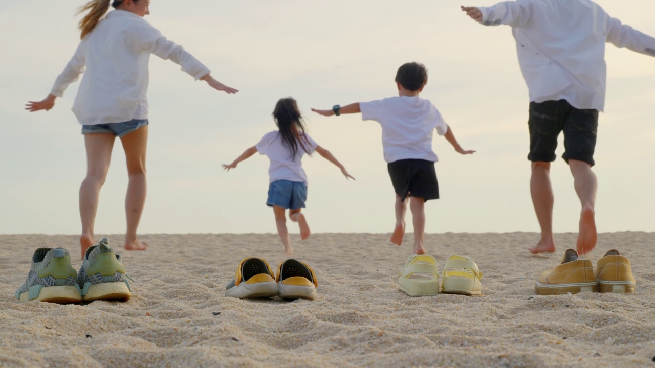 爸爸、妈妈和孩子脱鞋在沙滩上奔跑，背影的家庭，父母带着孩子手牵着手一起在沙滩上奔跑，在假期，旅行，积极的生活方式，快乐的家庭日视频下载