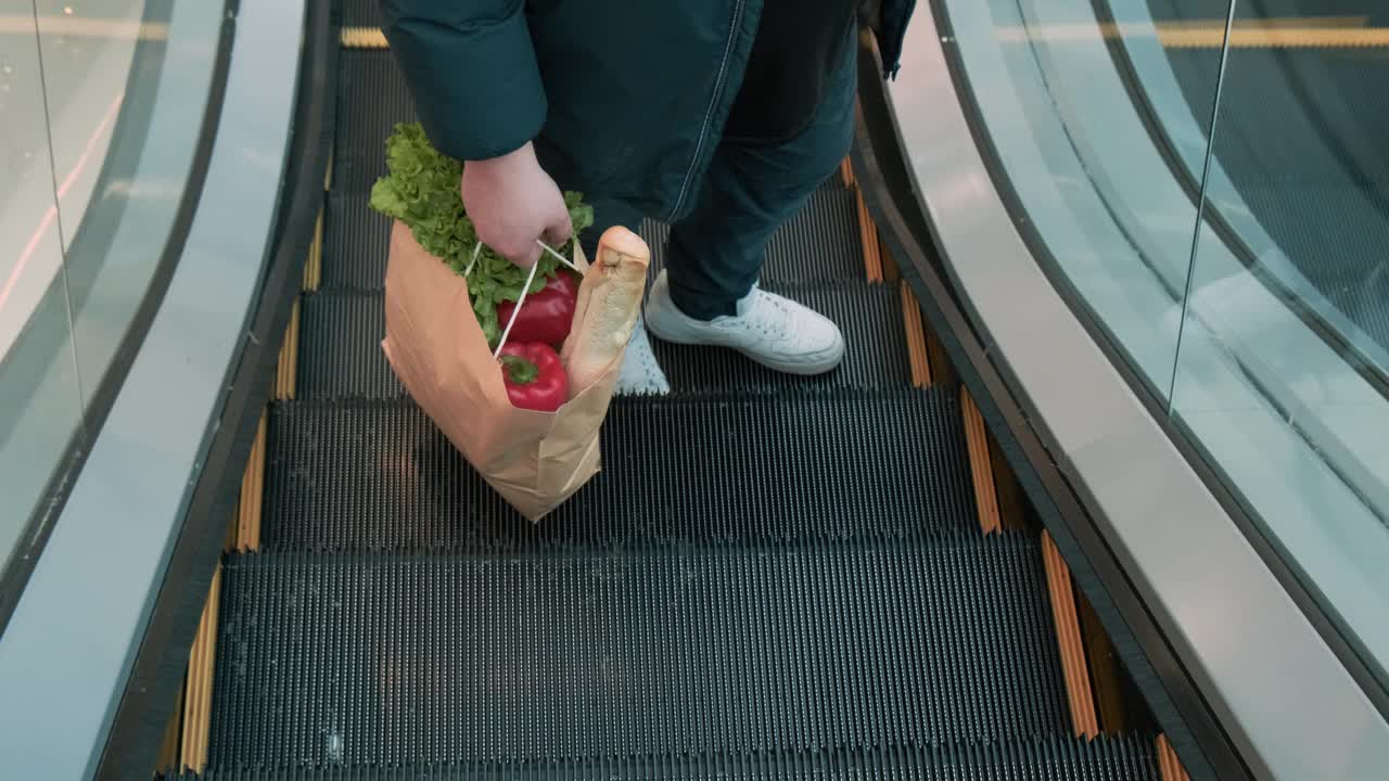 商场的自动扶梯上，一名男子手持装有素食食品的环保纸袋。可持续的生活方式。男性在超市购物视频素材