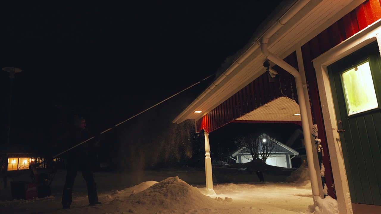 用长伸缩雪耙清除屋顶积雪，中年男子在天黑时用除雪工具工作。后侧视视频视频下载