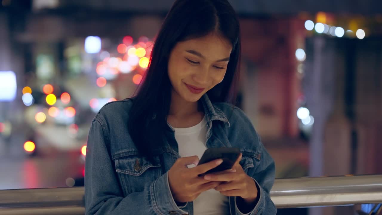 美丽的年轻亚洲女子站在城市街道在晚上使用手机。一个快乐的女人站在那里用手机打字视频素材